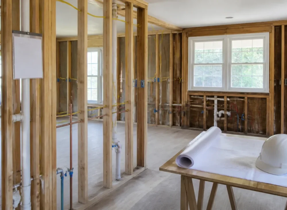 Kitchen remodel permit stage in a Massachusetts home, with open stud walls showing new wiring and plumbing, rolled plans and a hard hat ready for inspection